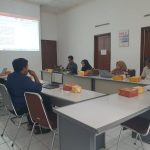 6 orang berkumpul mengeliling satu meja besar sedang menyimak dokumen yang ditampilkan di depan menggunakan proyektor.