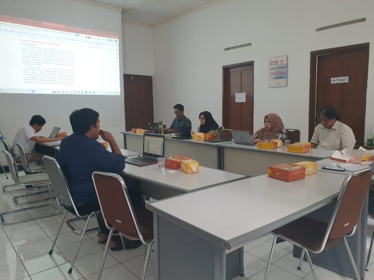 6 orang berkumpul mengeliling satu meja besar sedang menyimak dokumen yang ditampilkan di depan menggunakan proyektor.