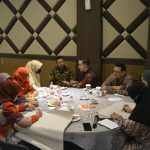 Gambar berisi 8 orang sedang mengelilingi meja bundar. Ada yang menyampaikan pendapat dan ada yang menulis.