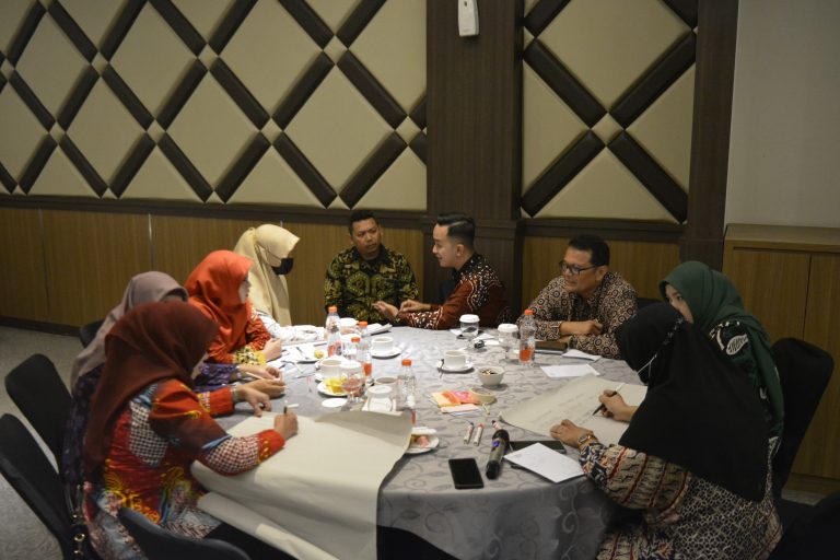 Gambar berisi 8 orang sedang mengelilingi meja bundar. Ada yang menyampaikan pendapat dan ada yang menulis.