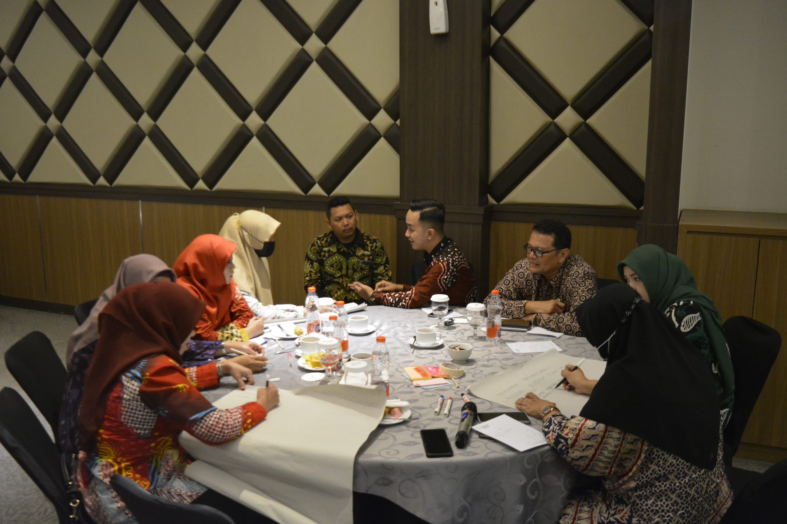 Gambar berisi 8 orang sedang mengelilingi meja bundar. Ada yang menyampaikan pendapat dan ada yang menulis.