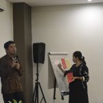 Gambar berisi dua orang sedang memfasilitas diskusi menggunakan pengeras suara dan papan flip chart.