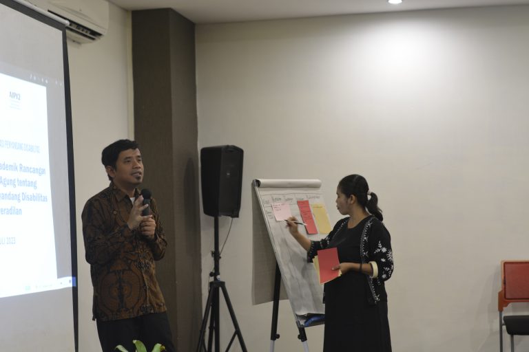 Gambar berisi dua orang sedang memfasilitas diskusi menggunakan pengeras suara dan papan flip chart.