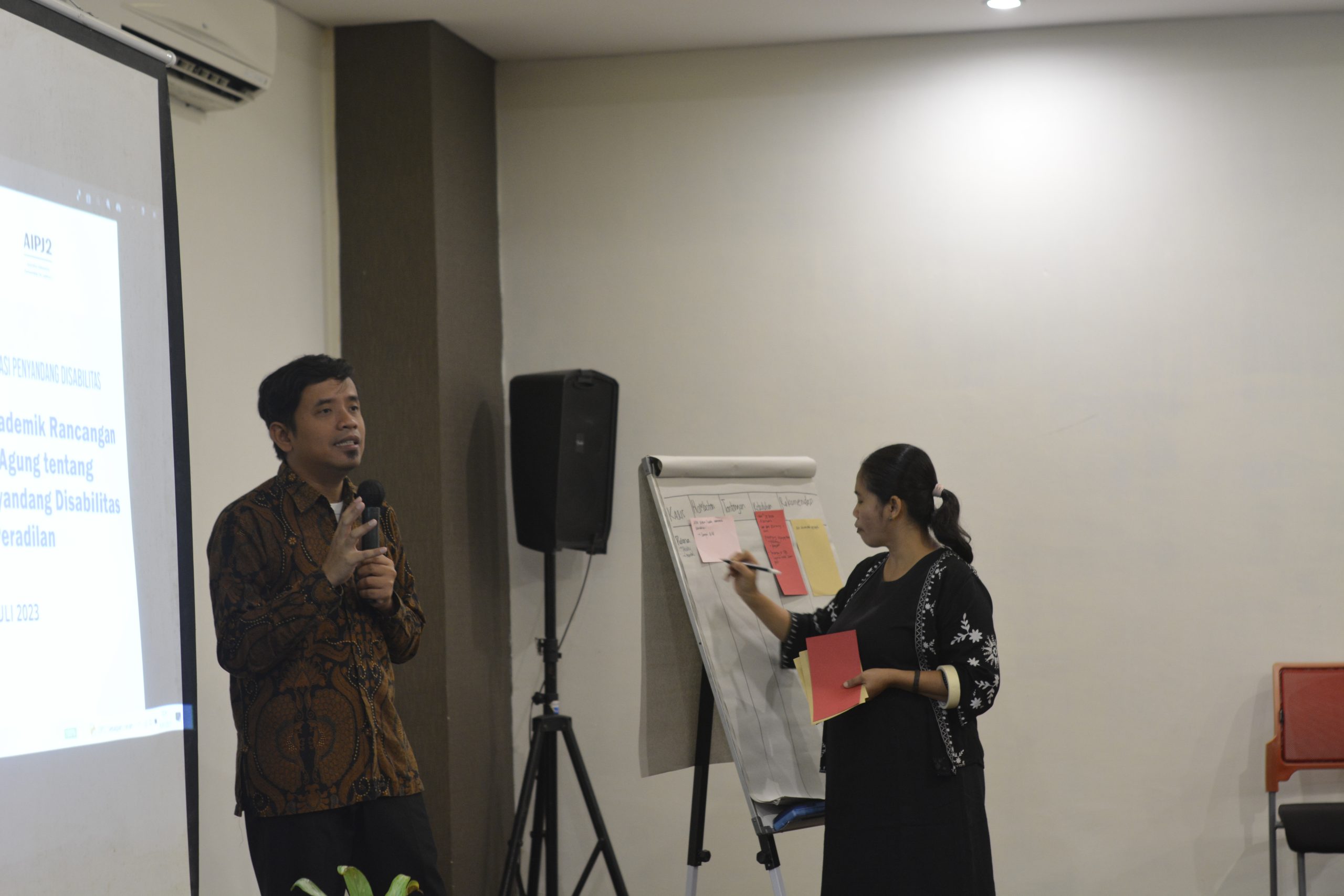 Gambar berisi dua orang sedang memfasilitas diskusi menggunakan pengeras suara dan papan flip chart.